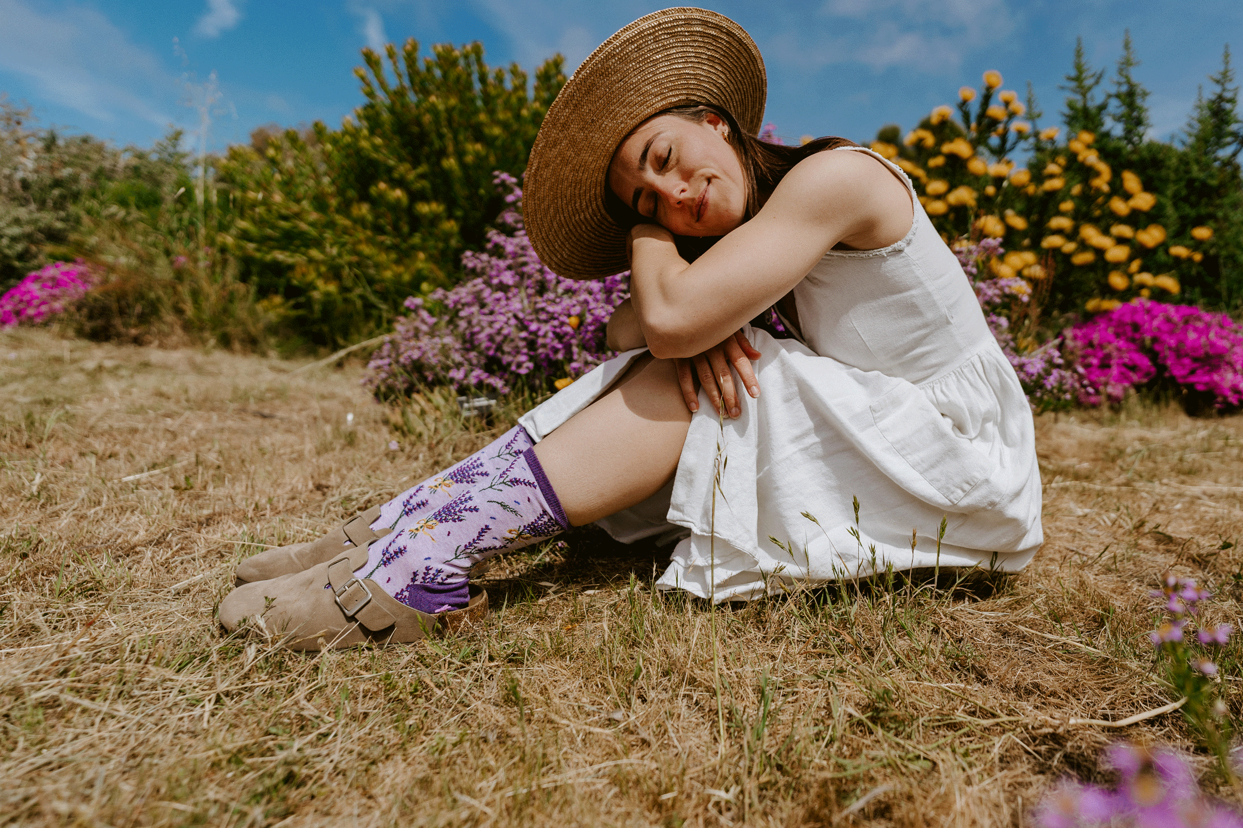 A woman sitting on dry grass in a field, wearing a white sleeveless dress, a wide-brim straw hat, and purple patterned socks, with flowers and greenery in the background.