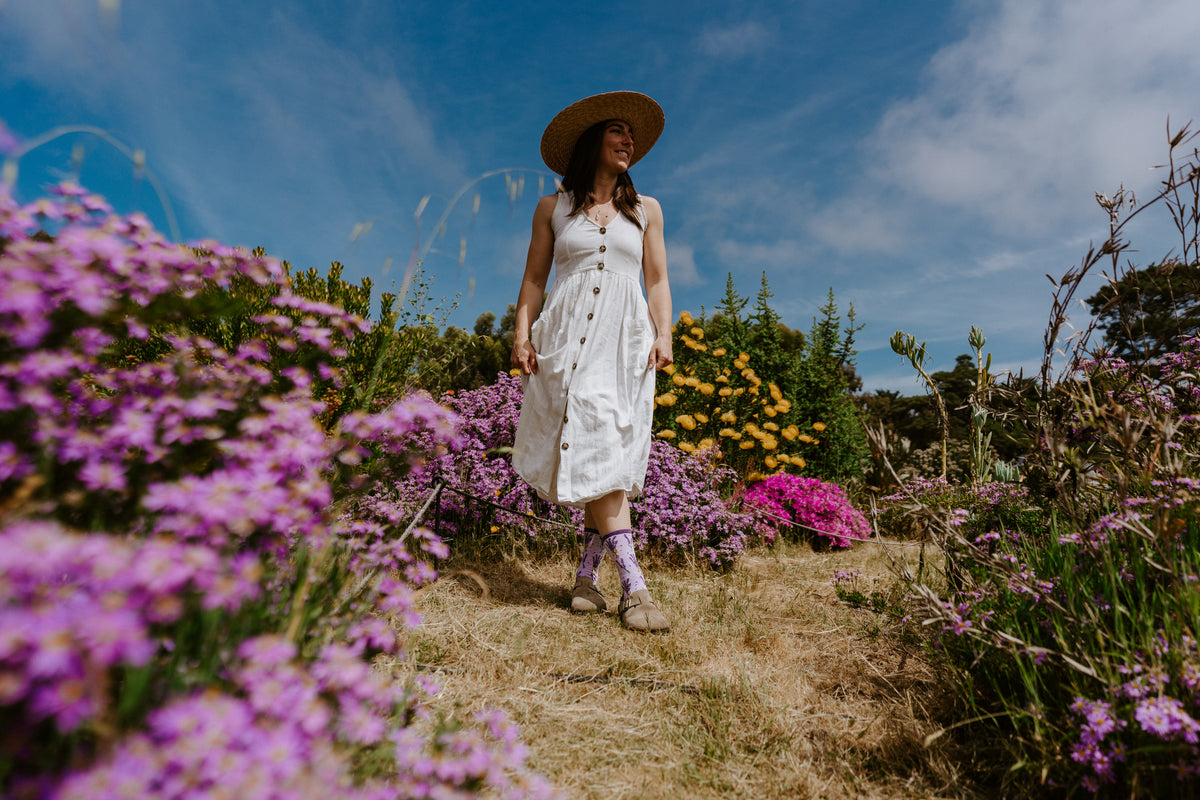 Woman walking in amongst purple wild flowers wearing lavender-themed socks with a white dress and sunhat