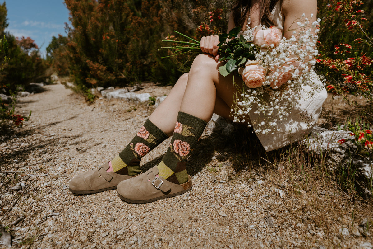 Close-up of a person sitting outdoors on a gravel path wearing dark olive green floral socks with roses on.