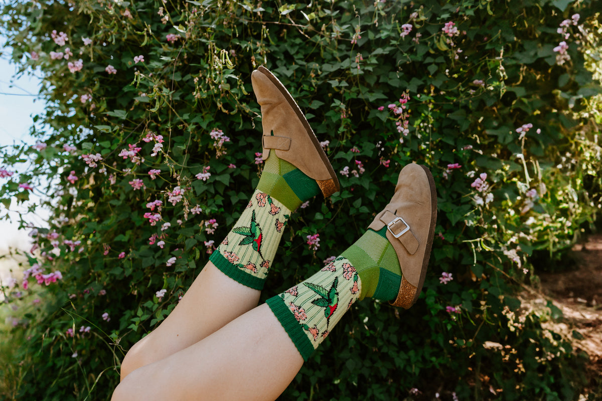 A person's legs are raised in front of a leafy green garden with pink flowers, wearing hummingbird Merino wool socks.