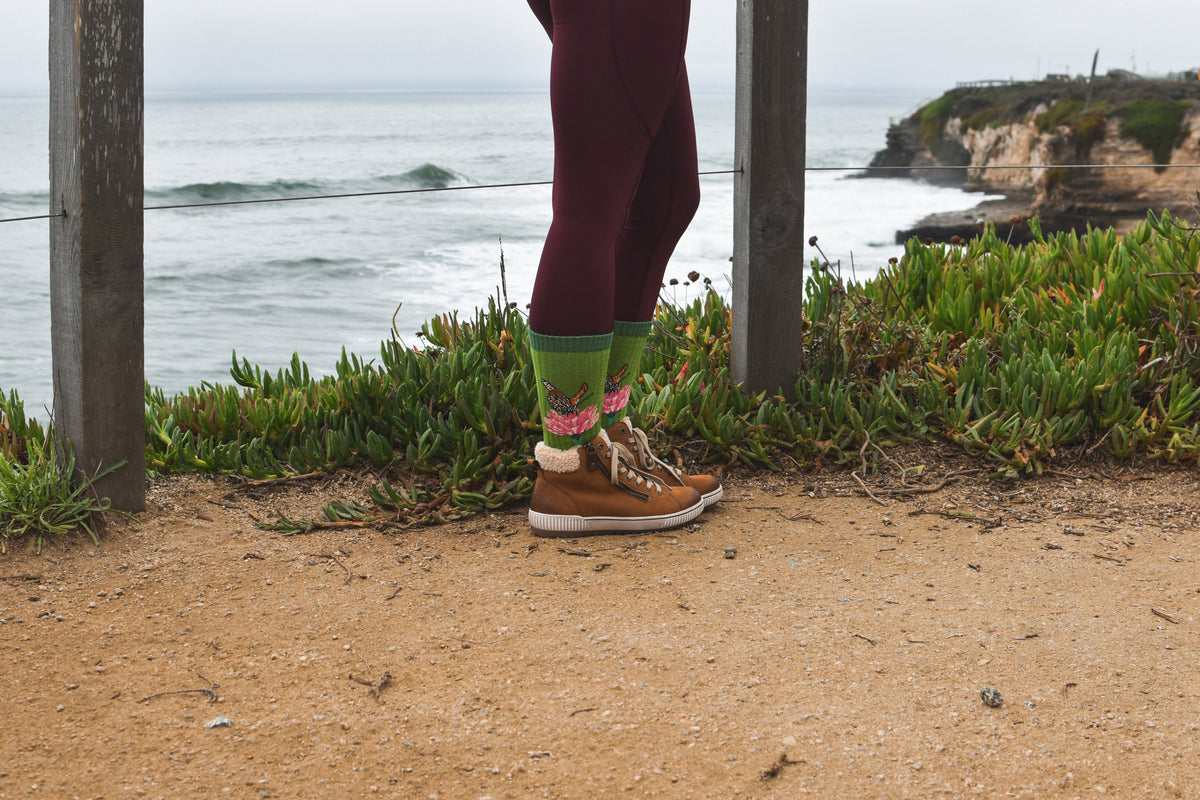 A person standing on a sandy coastal path with the ocean and cliffs in the background, wearing Merino wool socks with a butterfly and flower on.