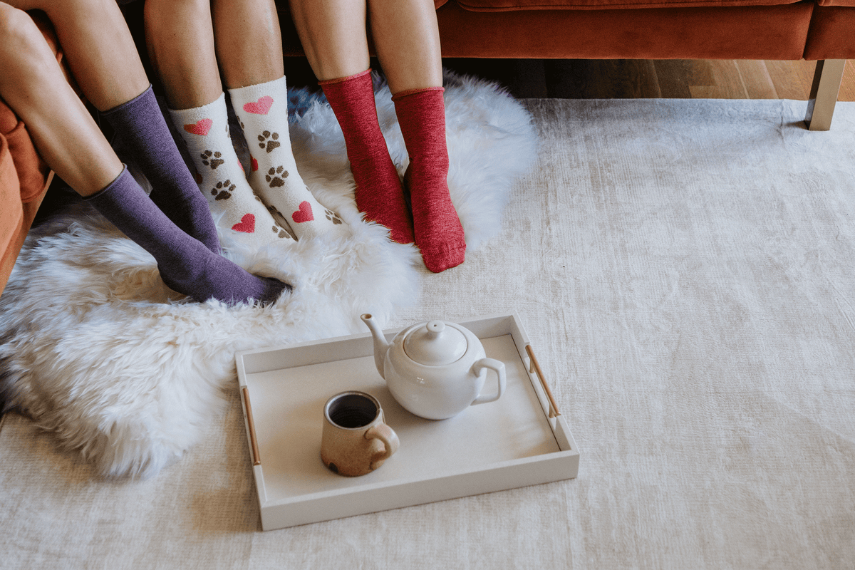Three pairs of cozy socks worn by people sitting on a white faux-fur rug on a light-colored floor, with a white tray holding a white teapot and a small cup in the foreground.